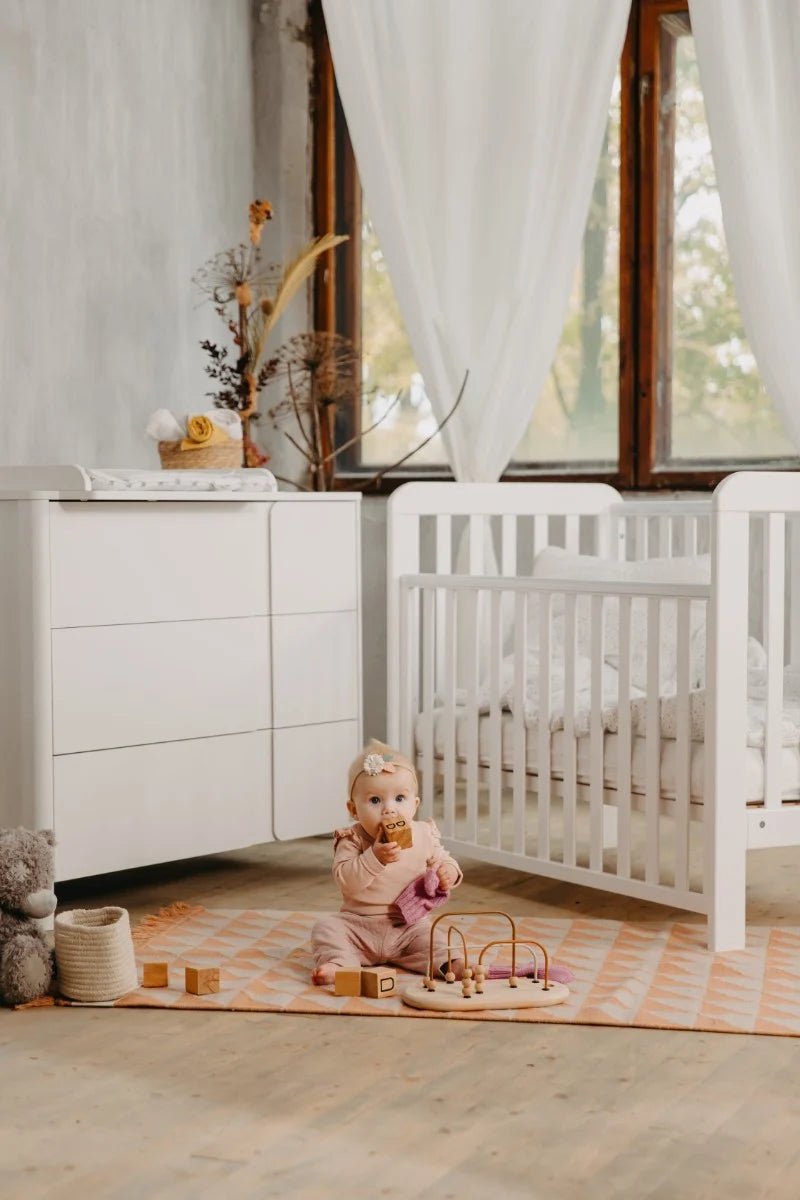 Sfeervol ingerichte kinderkamer met wit ledikant en commode, waar een baby speelt op een speelmat
